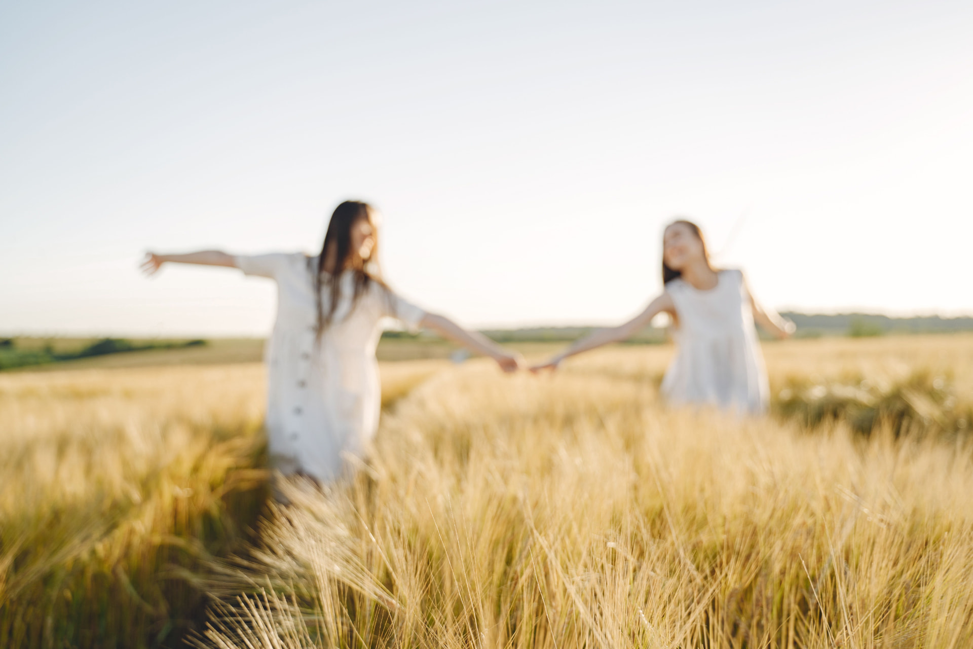 Two women in white dresses walking hand in hand through a sunlit wheat field
